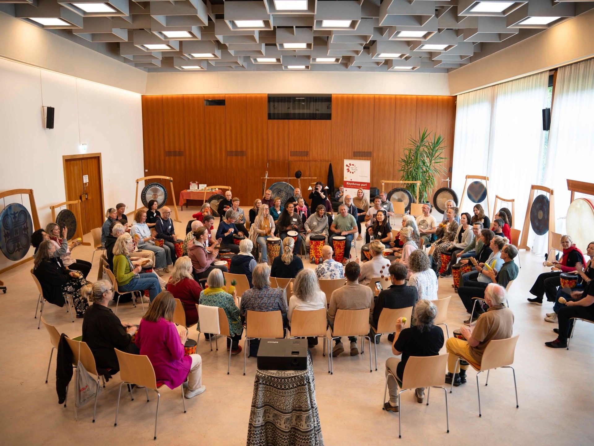 Großer Trommelkreis mit vielen Menschen in einem hellen Saal, Symposium 2026