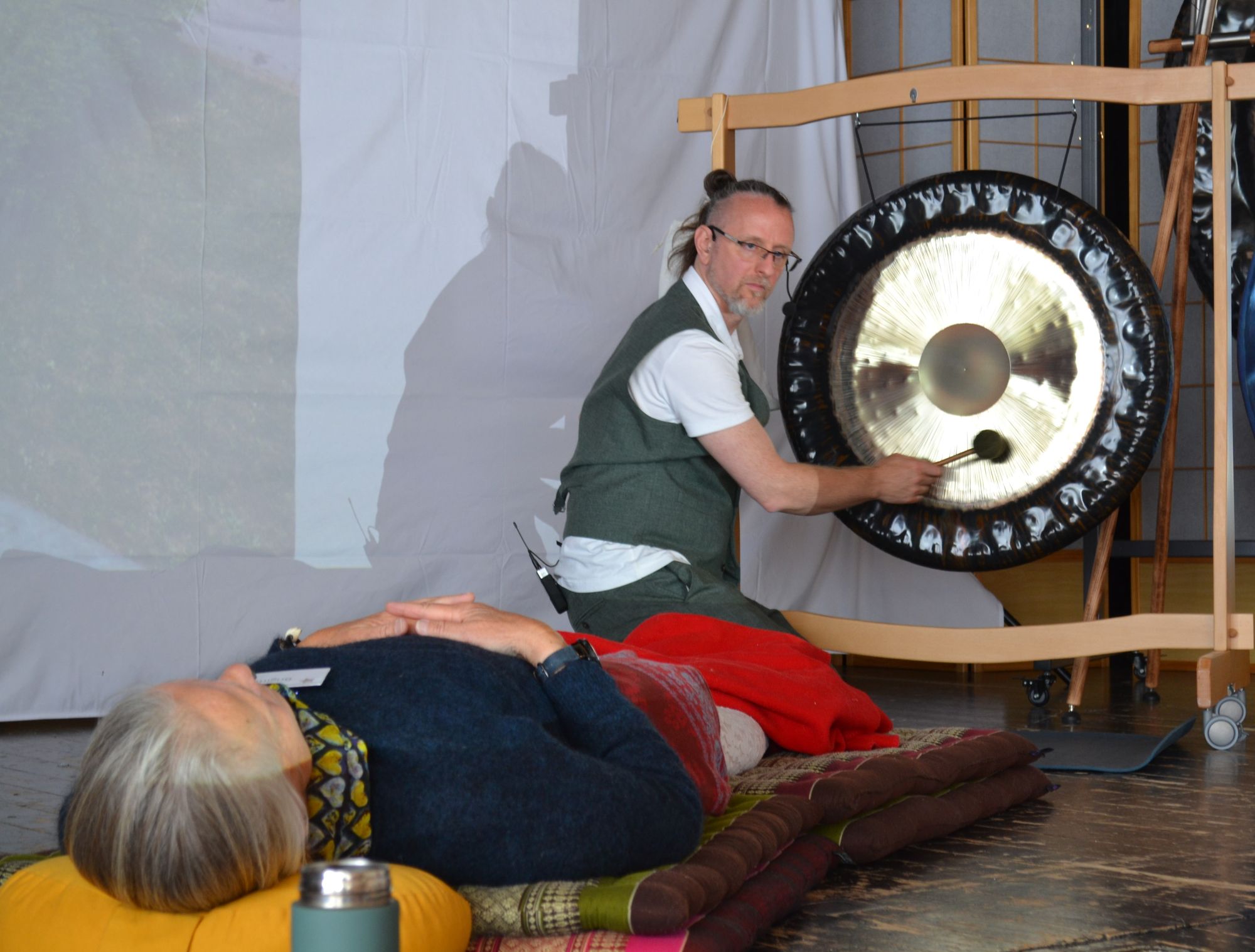Ein Mann spielt einen Gong neben einer liegenden Person, Symposium 2026