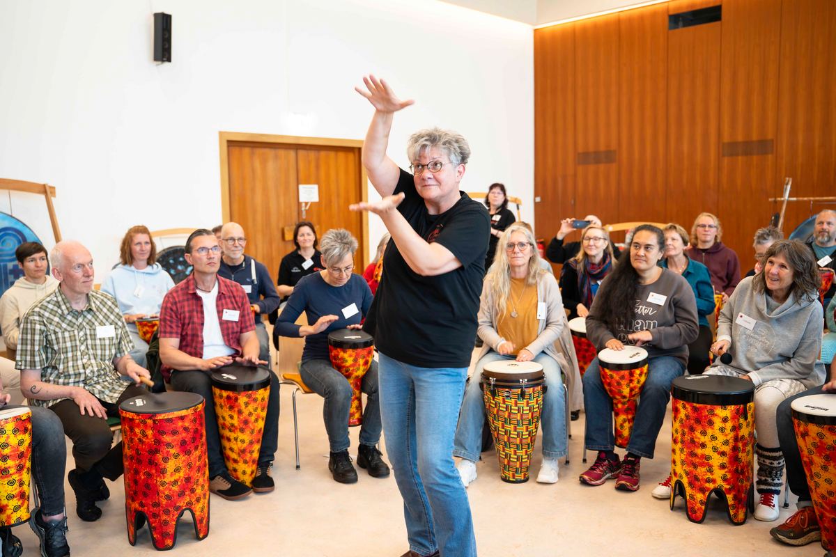 Lachende Menschen trommeln gemeinsam auf bunten Trommeln, Symposium 2026